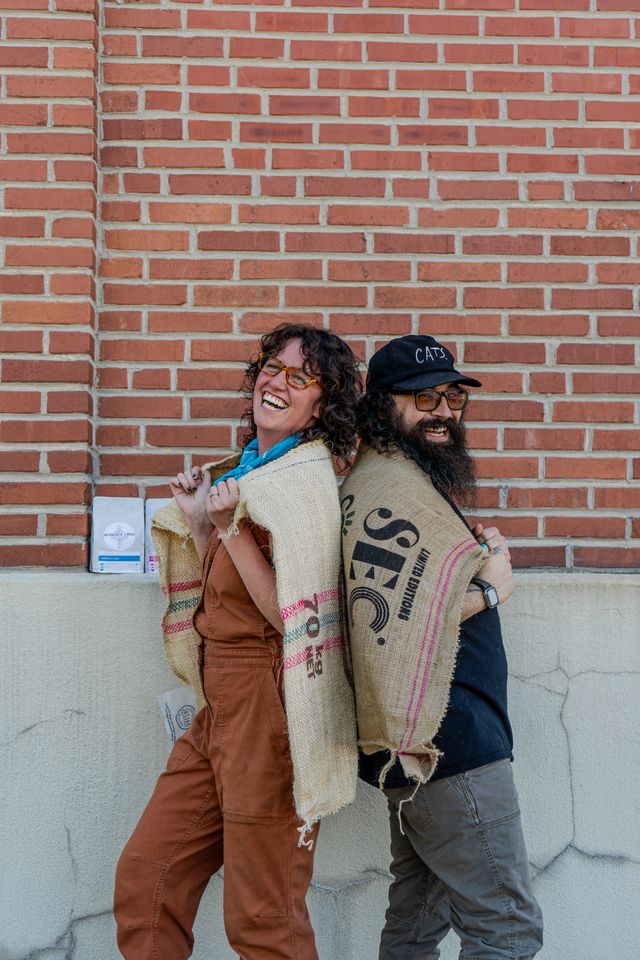 Two team members leaning back to back with burlap coffee sacks across their shoulders