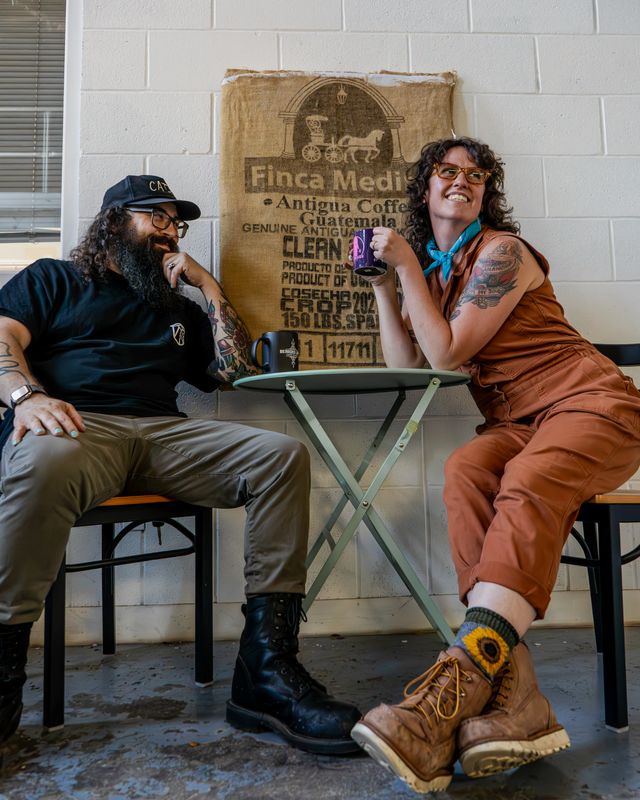 Two Bearded Lady team members laughing over mugs at a patio table in front of a burlap coffee sack