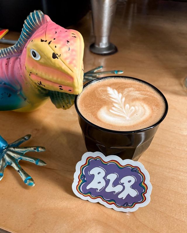 A latte with leaf art beside a colorful ceramic fish and a Bearded Lady sticker