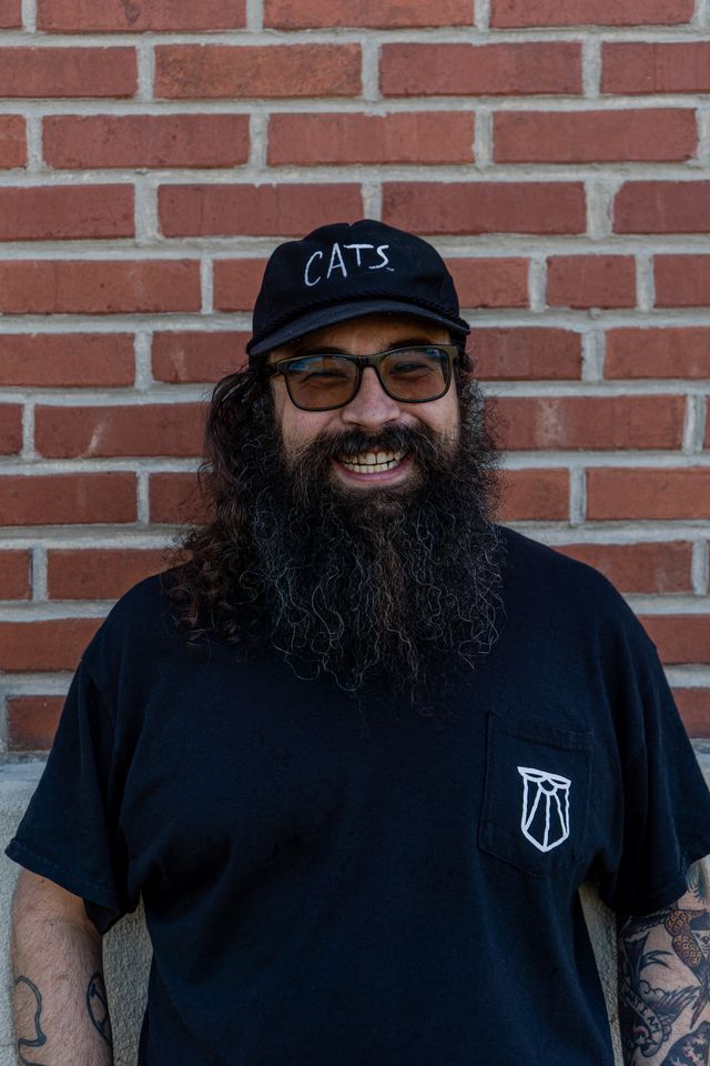 Team member with a full beard, glasses and a black cap, grinning against a brick wall