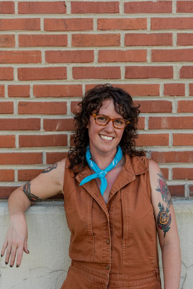 Team member in a rust jumpsuit and teal neckerchief leaning on a brick wall, smiling