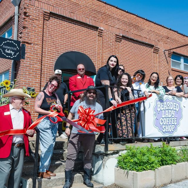 A crowd gathered on the brick building steps for a ribbon cutting, with a large red bow and a Bearded Lady Coffee Shop banner