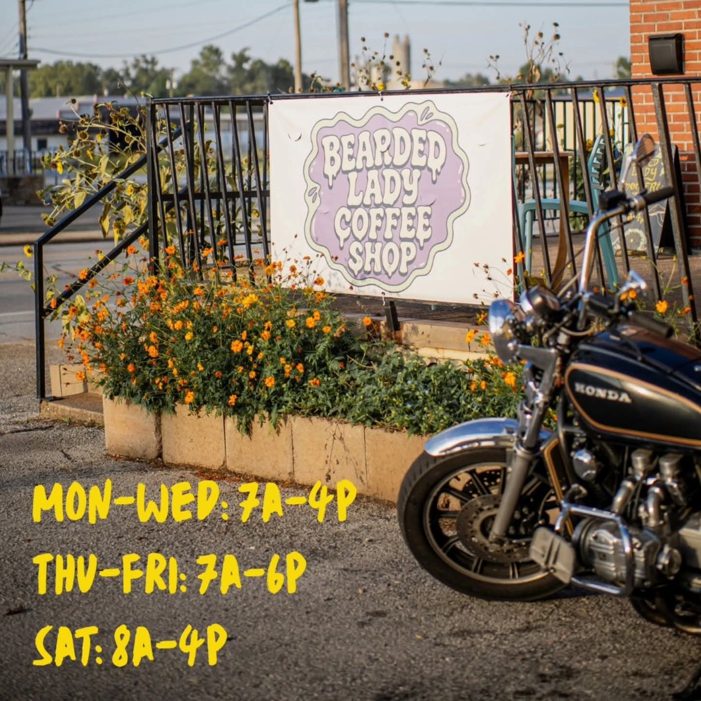 The Bearded Lady Coffee Shop sign among orange flowers, with a motorcycle parked beside it and the hours painted underneath